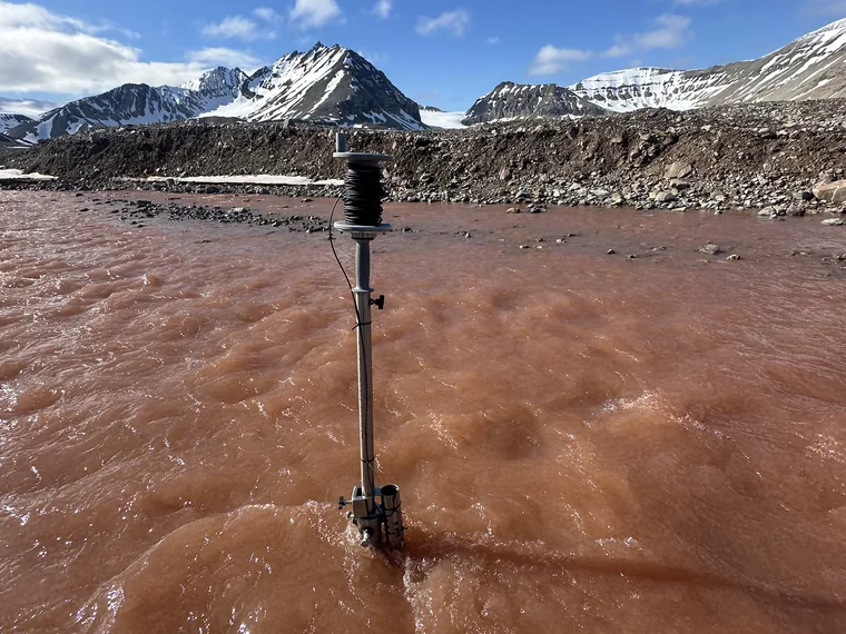 Palo con strumento di misura avvitato su una fondazione T-Block in mezzo al fiume glaciale, sotto un cielo azzurro e le vette innevate delle Svalbard.