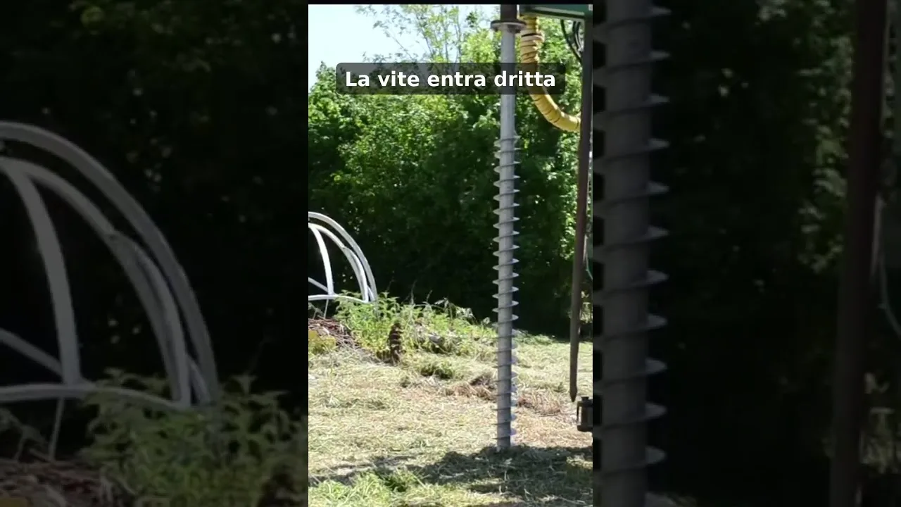 La macchina a colonna tiene in verticale una vite di fondazione T-Block, in un prato con strutture bianche sullo sfondo.