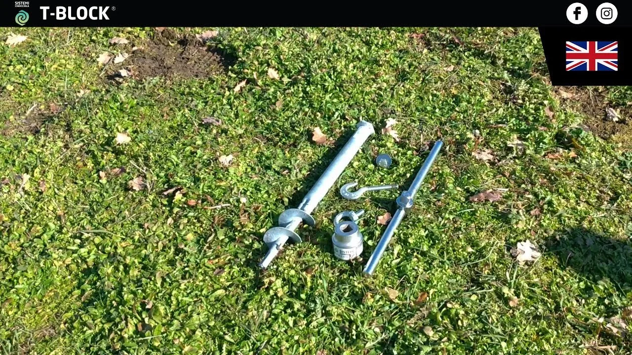 On a lawn, a 50 cm T-Block ground screw with a hexagonal head, the socket, the T-handle, a hook, an eyebolt and a washer.