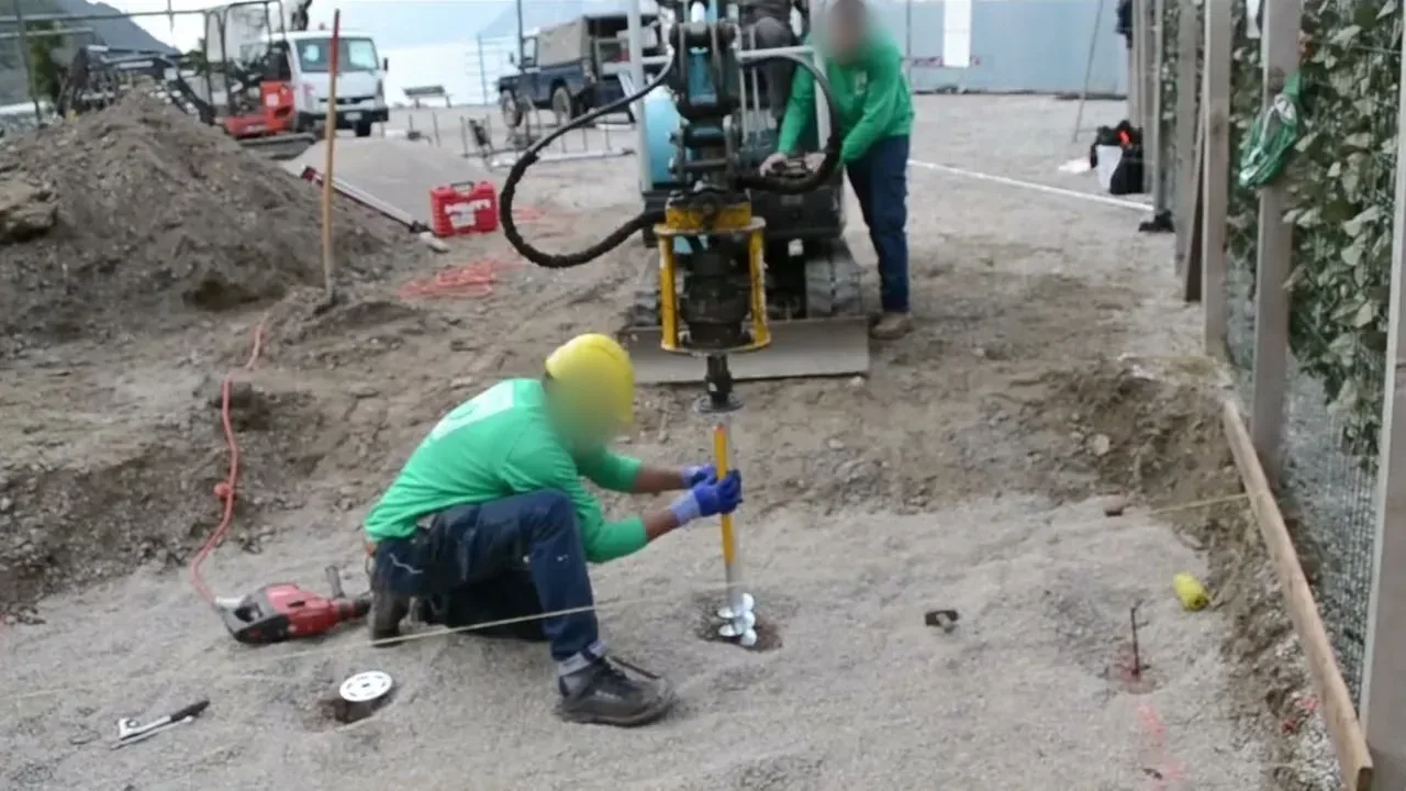 Cantiere a Torbole: un operatore tiene in verticale una fondazione a vite T-Block mentre il miniescavatore la avvita nella ghiaia.