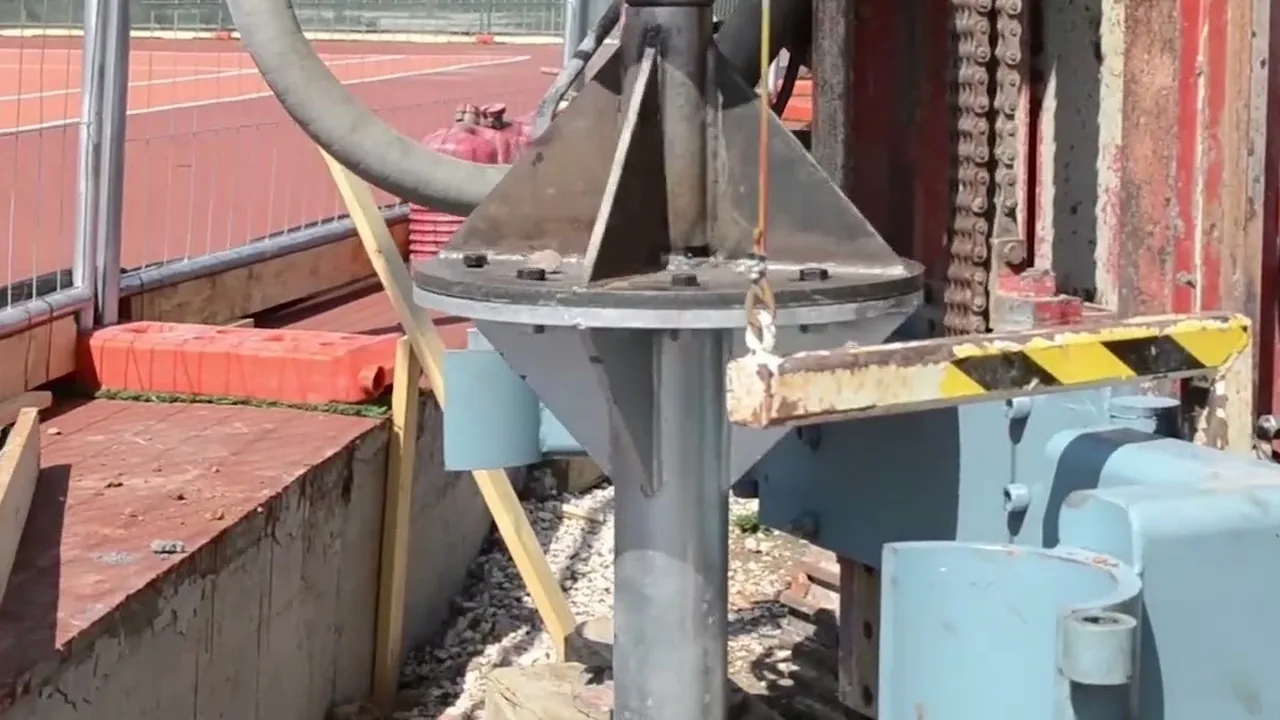 La testa a flangia di un T-Block da 3 metri agganciata alla macchina cingolata, accanto a un campo da tennis in terra rossa.